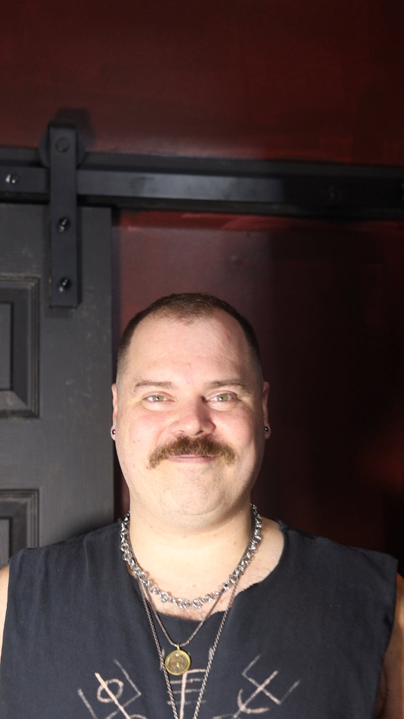 A man with a mustache wearing a black sleeveless shirt with white graphic symbols and layered silver necklaces, standing in front of a dark reddish-brown door with a black metal bracket. salem witch store
