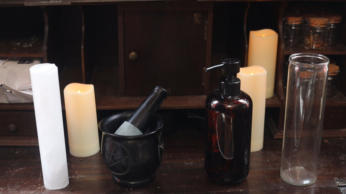 A collection of mystical and occult-themed glass containers on a dark wooden shelf, including a black pentagram mug, a dark amber pump bottle, a clear glass cylinder, and white pillar candles.