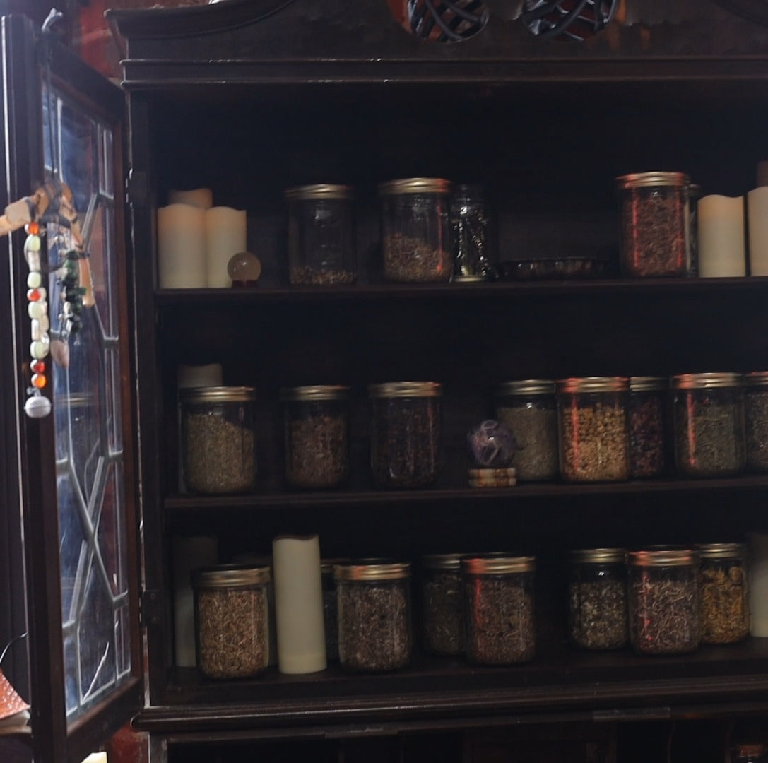 A dark wooden cabinet with three shelves displaying numerous glass jars filled with dried herbs or spices, alongside several white pillar candles. witchy herbs, dressing candles, candle making, spell candles