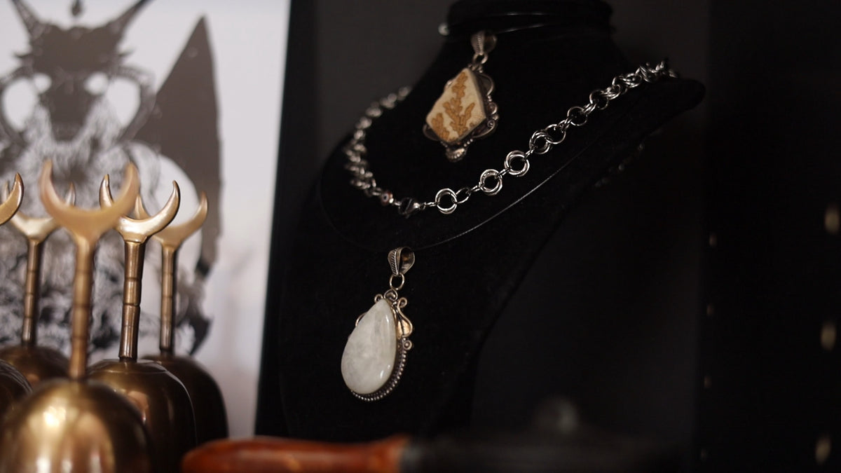 A silver-toned chain necklace displayed on a black mannequin bust, featuring a large white teardrop pendant and a smaller irregular beige stone pendant. The bust is photographed in a moody setting with decorative brass objects blurred in the foreground.