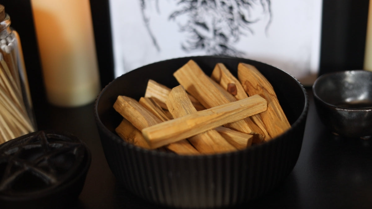 A black ribbed bowl filled with natural wood incense sticks, set on a dark tabletop with a blurred candle and decorative background. saging and cleansing, palo santo, smudging