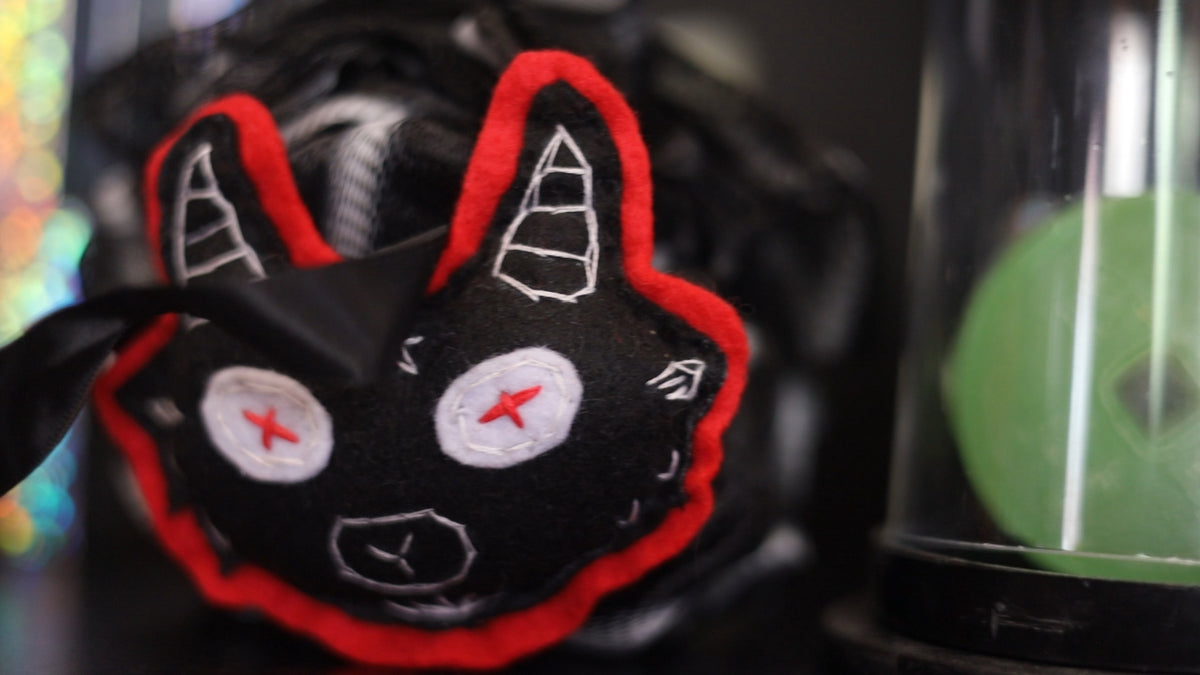 A close-up of a black felt monster face patch with red stitched border, white embroidered eyes and ears, and red stitched star eyes, shown against a dark blurred background.