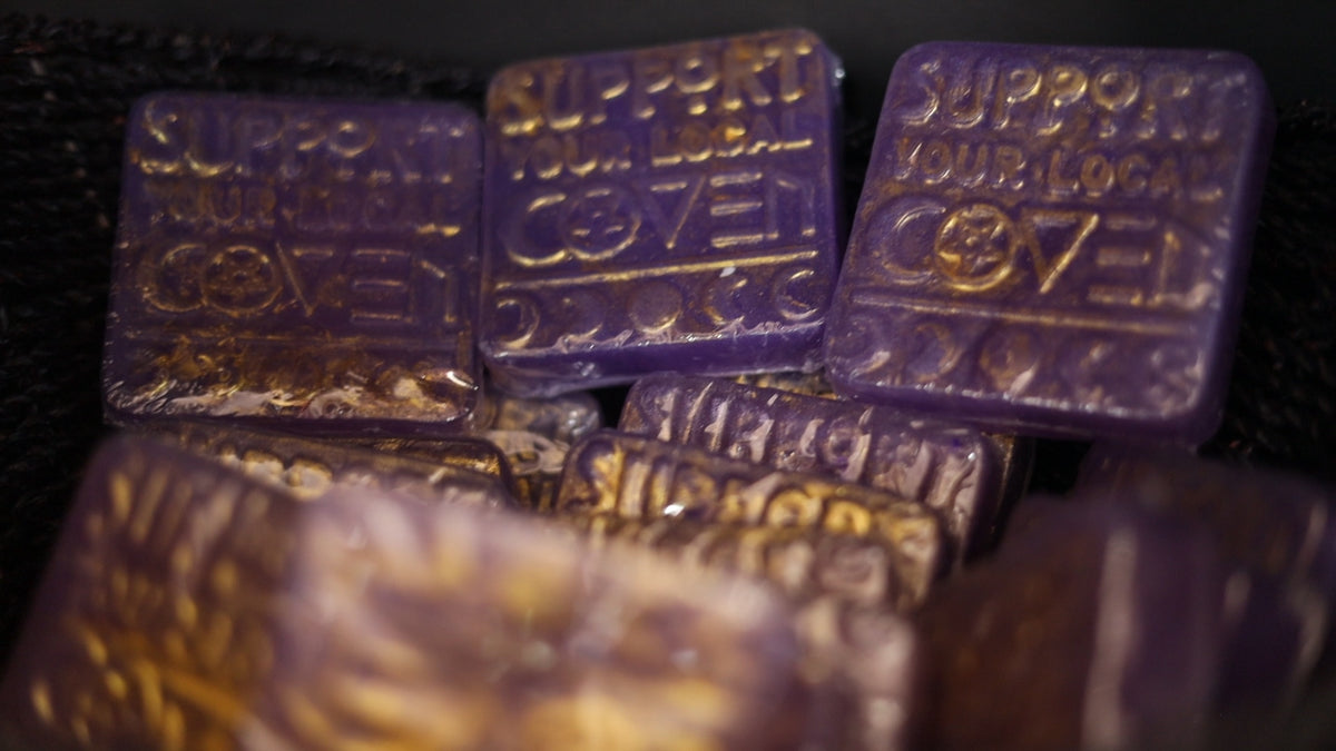 A close-up shot of several purple, translucent square wax melts stacked together on a dark background. The top wax melts are embossed with the text 'SUPPORT YOUR LOCAL COVENANT'. coven , witch coven, soap, handmade local soaps