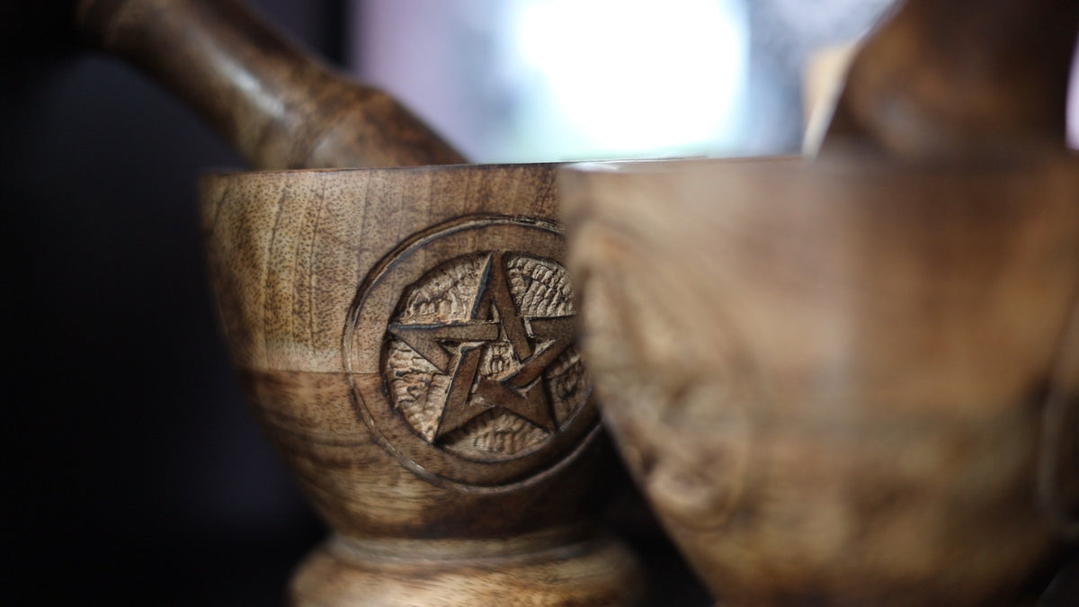 Close-up of a carved wooden mortar and pestle featuring an engraved pentagram symbol, with another wooden bowl partially visible in the foreground.