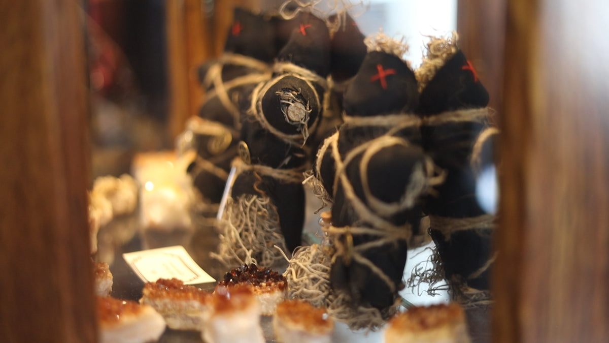 Three black smudge bundles wrapped in natural twine, with one featuring a white owl emblem and another with a red cross, displayed in a wooden case. poppets, crystals 