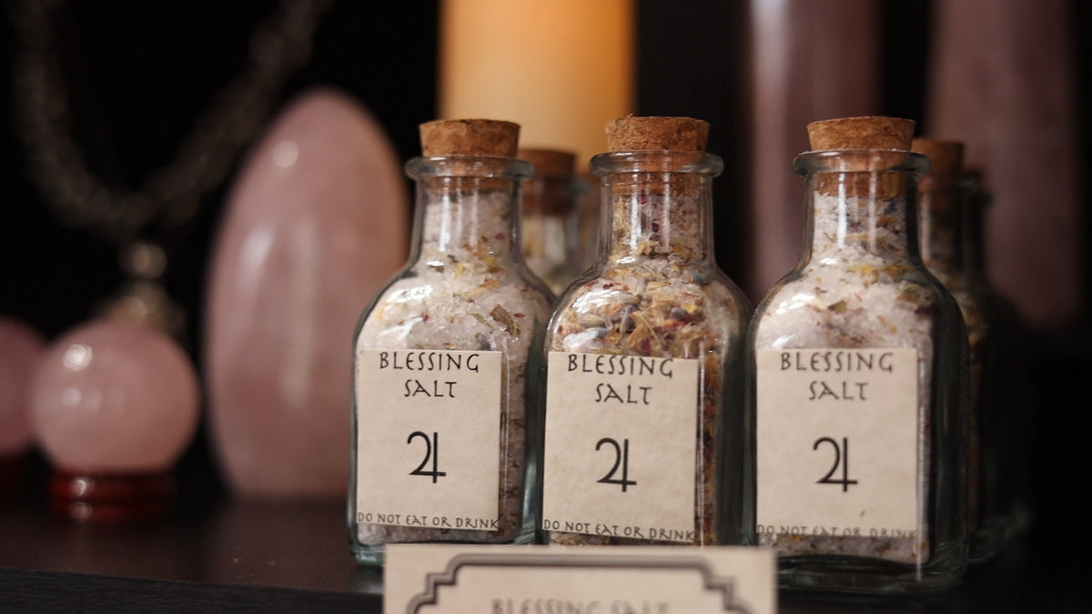 Three small glass bottles with cork stoppers labeled 'Blessing Salt 24' filled with salt crystals, arranged on a dark surface with blurred pink stones and candles in the background.