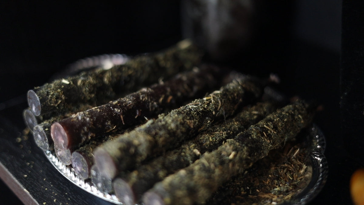 Several dark, herb-coated cylindrical sticks arranged on a silver tray against a very dark background. candles with herbs
