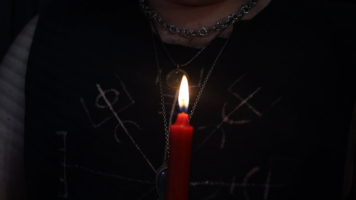 A close-up of a person wearing a black shirt with occult-style symbols, layered silver necklaces, and holding a lit red candle in a dark setting.